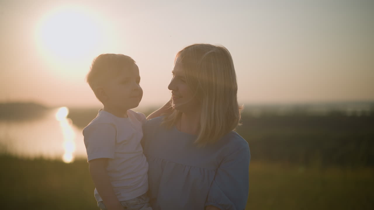 A heartwarming moment between a mother and her young son in a grassy field during sunset. The woman, dressed in a blue gown, lovingly holds her son in a white shirt as they share a tender smile