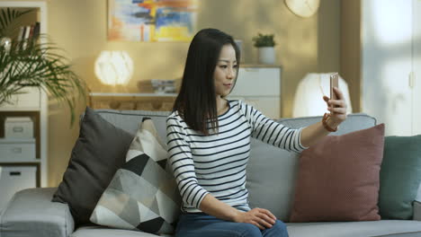 Mujer-Joven-Con-Pantalones-A-Rayas-Posando-Para-La-Cámara-Del-Teléfono-Inteligente-Y-Sonriendo-Mientras-Tomaba-Fotos-Selfie-Sentada-En-El-Sofá-En-La-Sala-De-Estar