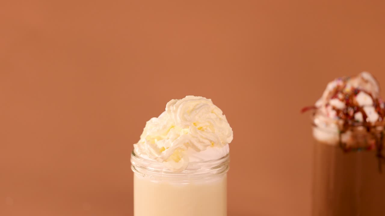 Vanilla milkshake topped with whipped cream, surrounded by cookies and chocolate, under warm studio lighting