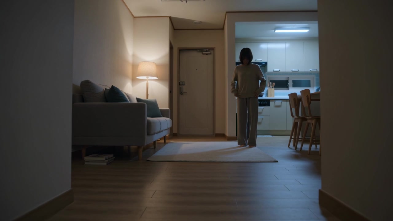 Donna in abbigliamento casual da casa che si alza e attraversa il tappeto verso il bancone della cucina, con una lampada accesa.
