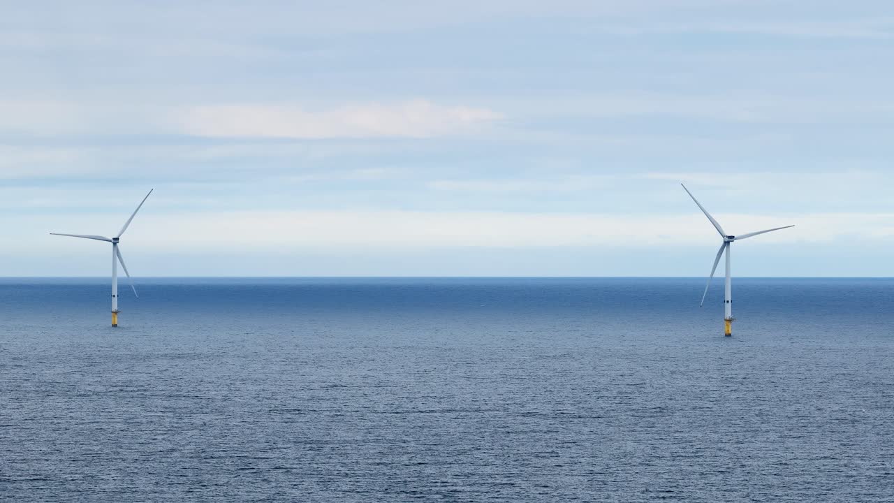 Two offshore wind turbines slowly rotate above a tranquil sea under soft daylight, captured in a steady wide shot with a serene, minimalist mood