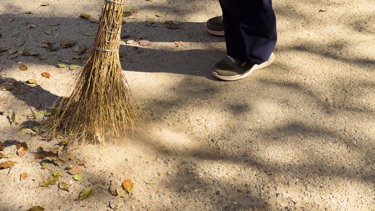 Nærbilde av en arbeider som feier tørre blader på en solfylt sti i Kyoto
