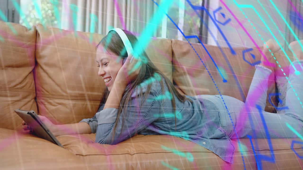 Teen girl lying on sofa fixing headset and tapping tablet causing neon overlays for entertainment