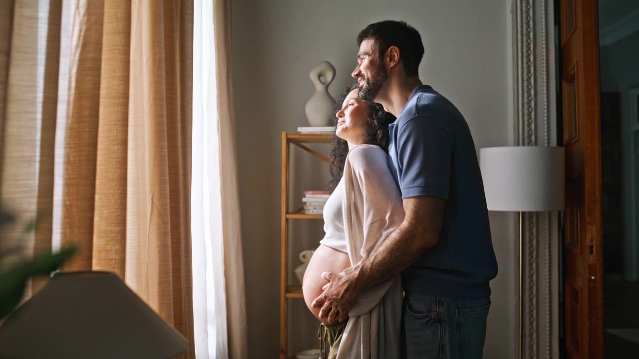 Expectant parents embracing near window. Man kissing pregnant wife forehead