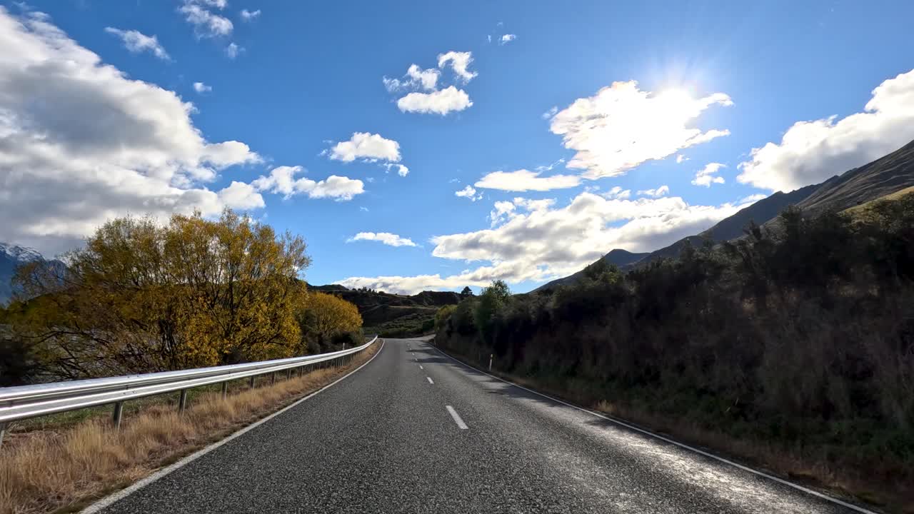 Point-of-view drive beside Lake Wakatipu, sunlit mountains, blue sky, and autumn foliage visible