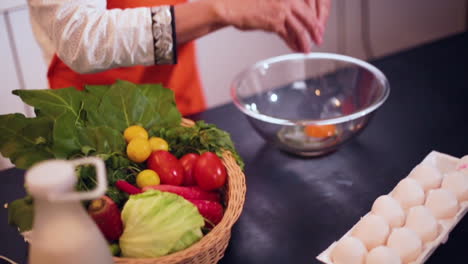 a slow mo close up view from top of a lady breaking an egg in the bowl in the kitchen, a basket of veggies with carrots, tomatoes and garlic on the table, the chef wore and orange colored apron