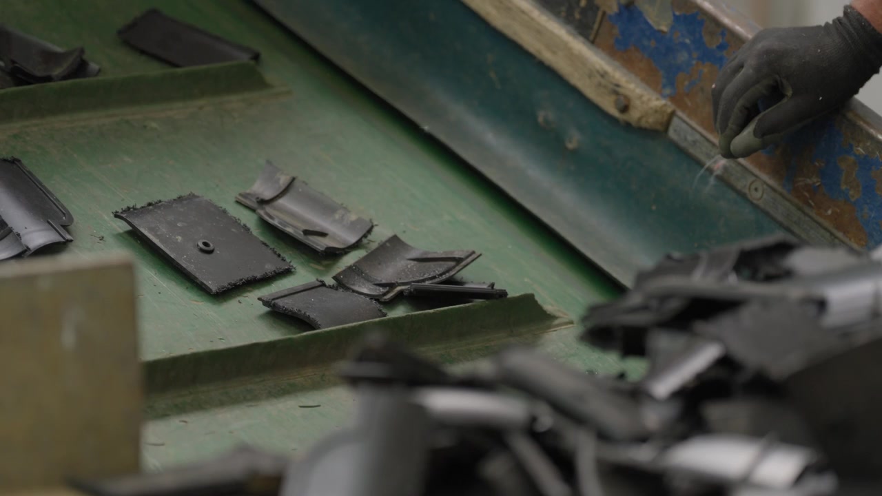 Gloved worker hand sorts fragments of shredded black plastic on worn green factory work surface