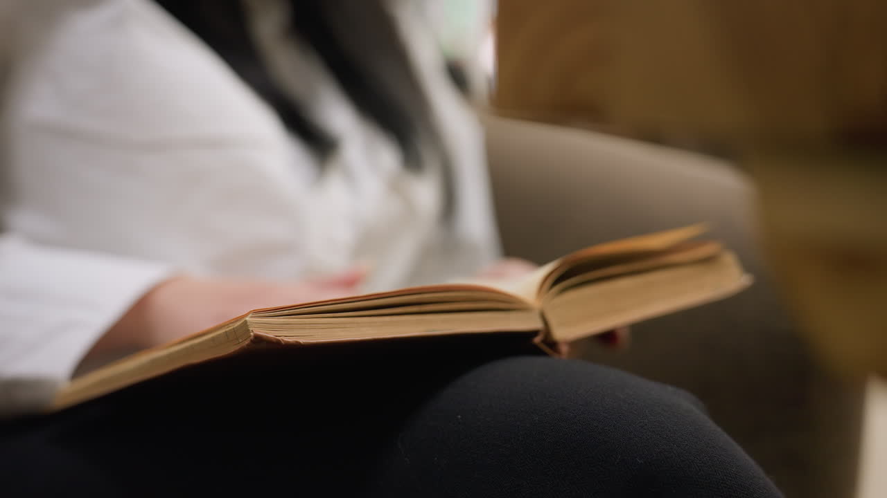 lady seated on soft chair flipping book page with relaxed posture and focused gesture as vintage book rests on leg, cozy indoor setting with blurred background