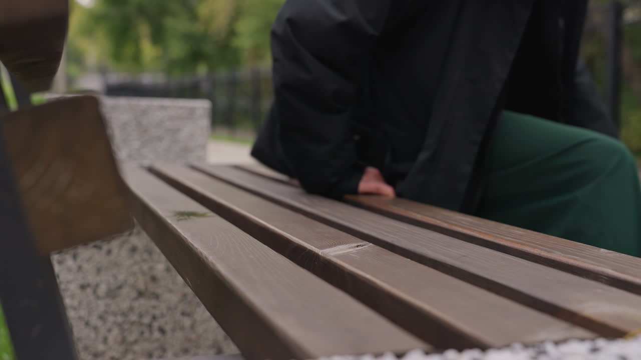 Parkbænk tæt på, person nærmer sig. Ærlige optagelser viser en person, der går hen mod en træbænk på en grussti, en hånd rækker ud efter kanten af sædet, iført grønne bukser og mørk frakke, jerngitter.