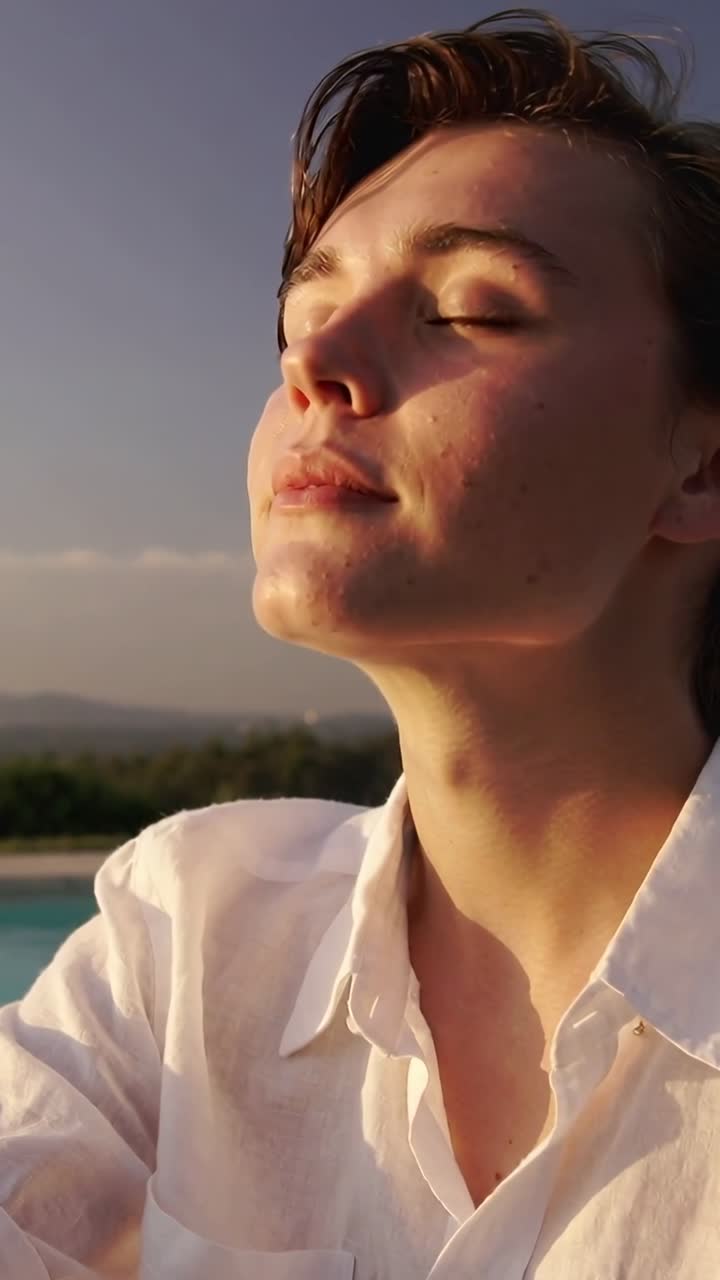 Video verticale: Uomo con camicia bianca che inclina la testa esponendo il viso al sole che gli riscalda la pelle a bordo piscina.