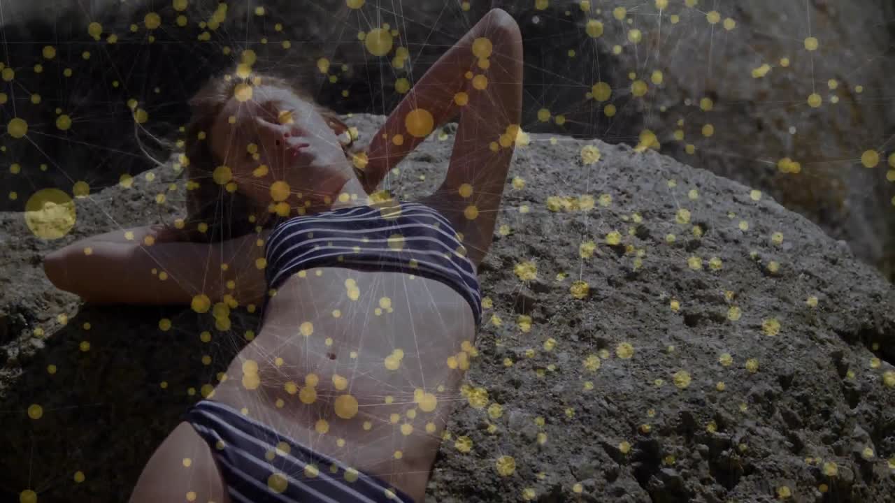 Modella sdraiata su una roccia che indossa un costume a righe, in posa tra un effetto di particelle dorate per la moda