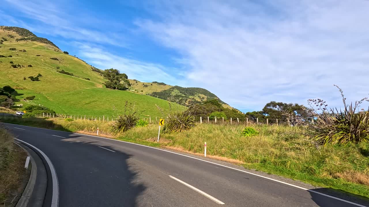 A vehicle travels along a winding rural road through lush green hills and farmland under bright daylight, captured in smooth, fast-motion timelapse style