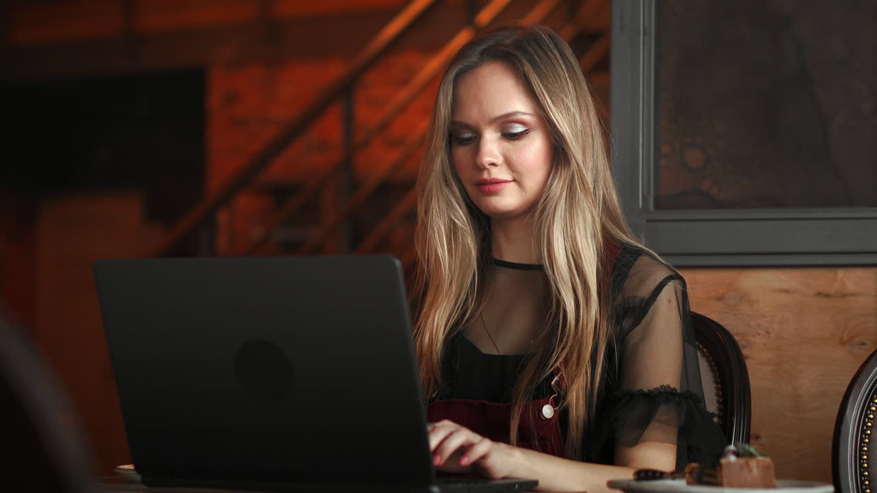 Young woman sitting in coffee shop at wooden table, drinking coffee and using smartphone.On table is laptop. Girl browsing internet, chatting, blogging. Female holding phone and looking on his screen.