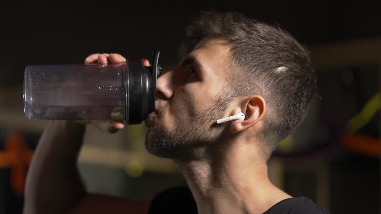Uomo robusto che beve acqua dalla bottiglia in una palestra buia, primo piano.