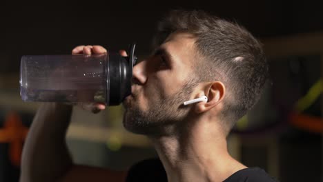 Tough-man-drinking-water-from-the-bottle-in-the-dark-gym,-close-up