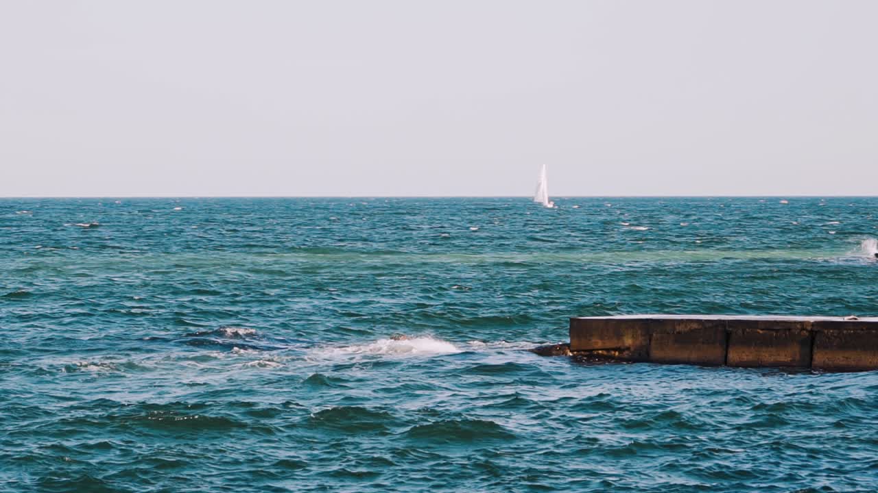 Motion of sea water. Waves break at stone pier and make white foam. Beautiful blue sea and white sail floating on alone. Slow motion.