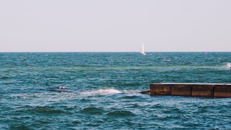 motion of sea water. waves break at stone pier and make white foam. beautiful blue sea and white sail floating on alone. slow motion.