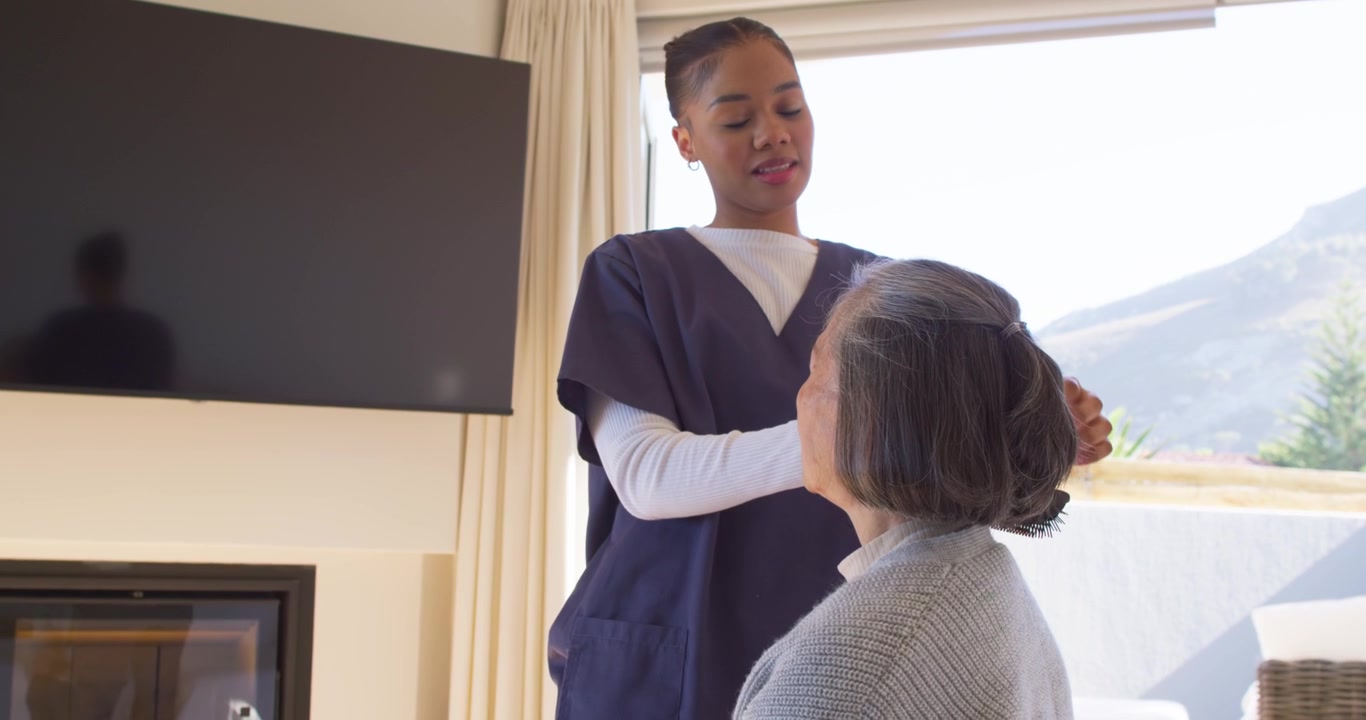 African American aide scrub-clad greeting styling Asian senior into bun for care with comb at home
