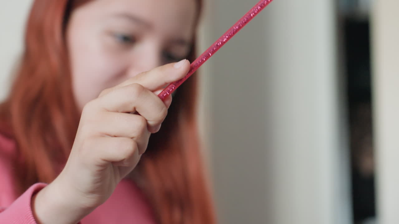 Girl Tests Magic Tool, Aspiring Female Magician Closely Observes Pink Prop Under Warm Indoor Lighting, Young Redheaded Girl Thoughtfully Examines Pink Wand While Testing Her Magic Skills Indoors