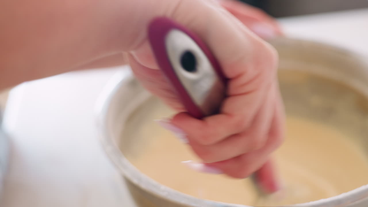 Close up of baking process as woman stirs smooth batter in stainless bowl with whisk, highlighting hand movement and texture during breakfast preparation in bright kitchen environment