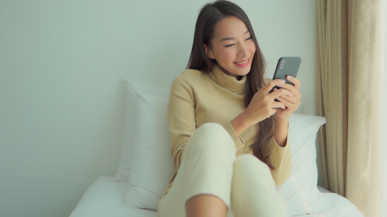 Smiling beautiful asian woman chatting on smartphone on bed
