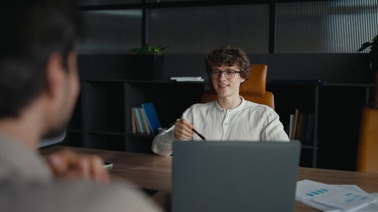 Over the shoulder a happy young guy with curly hair wearing glasses in a white shirt is having an interview with an experienced employee in the office