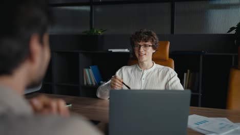 Over-the-shoulder-a-happy-young-guy-with-curly-hair-wearing-glasses-in-a-white-shirt-is-having-an-interview-with-an-experienced-employee-in-the-office