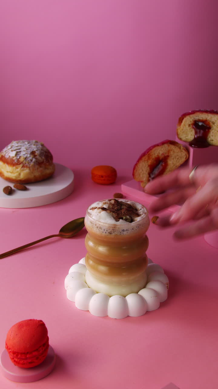 Mão gira delicadamente um latte em camadas com espuma cremosa em um copo escultural, cercado por donuts, recheio de geleia e adereços de estilização de alimentos em tons pastel sobre um fundo rosa para branding de café ou anúncios sociais
