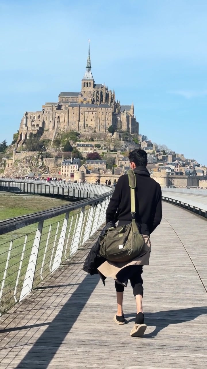 Uomo con borsone cammina su una passerella verso Mont Saint-Michel in una giornata di sole