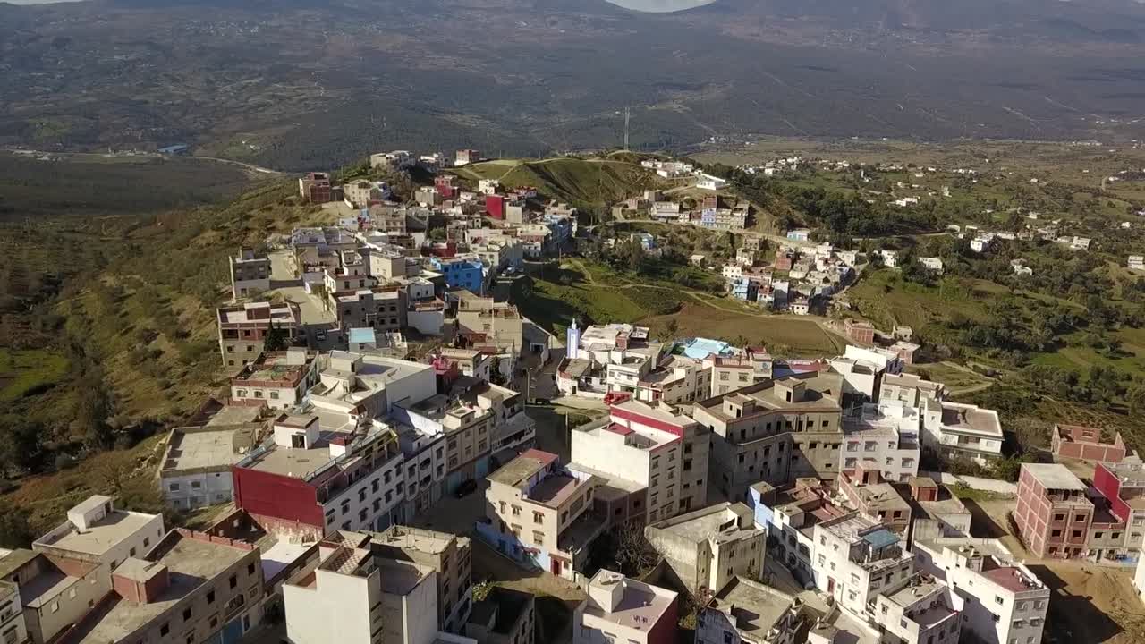 Splendida ripresa aerea ampia in HD di un tradizionale villaggio collinare nelle montagne del Rif vicino a Chefchaouen. Cattura l'architettura rurale colorata e il vasto paesaggio verde sotto un cielo sereno. Perfetto per i viaggi.