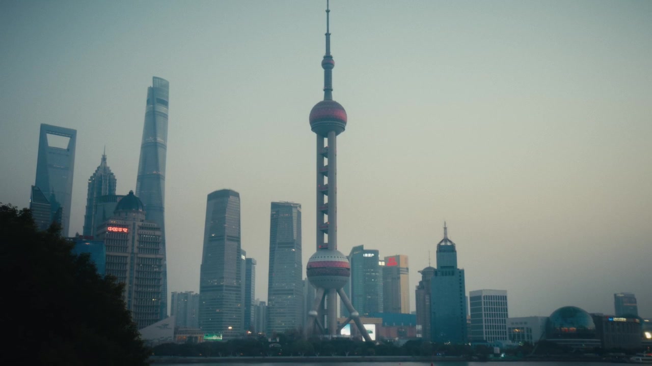 Lo skyline di Pudong a Shanghai è immerso nella foschia crepuscolare, con la Oriental Pearl Tower, lo Shanghai World Financial Center e la Jin Mao Tower che formano una silhouette drammatica contro il cielo serale
