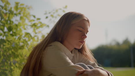 girl sits on grassy ground, appearing deep in thought with arms folded over her knees and head gently resting on her hand, with a blurred background of greenery, poles, and a distant stadium