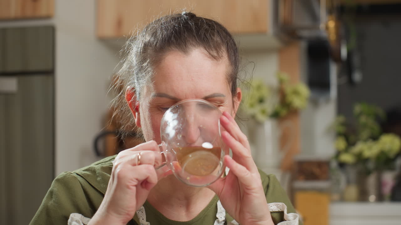 House wife drinks coffee from transparent glass cup with eyes closed, then drops cup gently while gazing at camera with calm satisfied expression in cozy kitchen