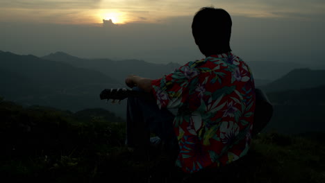 guitar player looks at the sunset with sunglasses at mountain top southeast asia wearing vintage colorful shirt