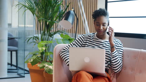 young adult mixed race businesswoman sitting on a sofa in a casual office using phone while working on a laptop computer balanced on her knees