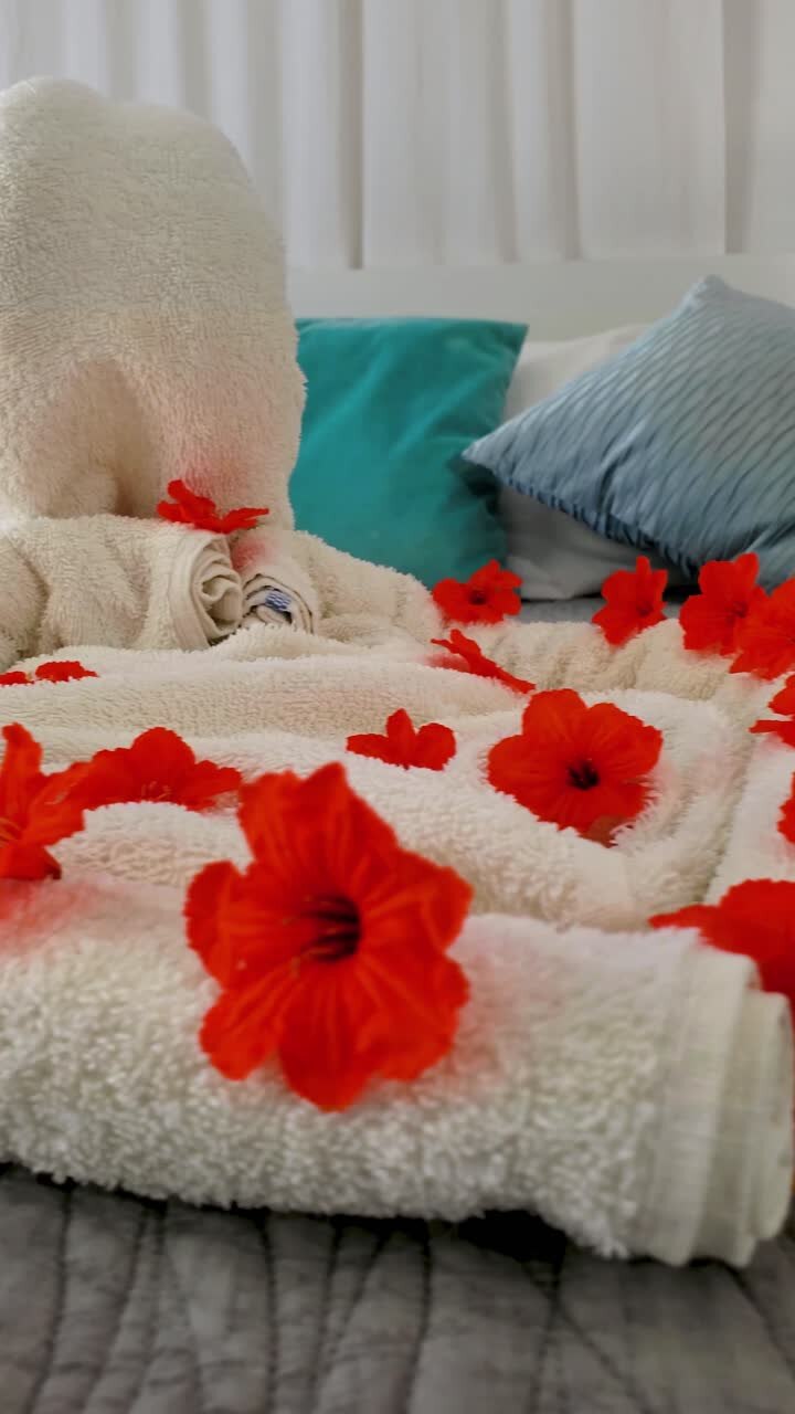 Fiori di ibisco rosso-arancio brillante con asciugamani arrotolati e piegati in forme decorative sul letto. Gesto di benvenuto in un ambiente alberghiero, con cuscini turchesi e grigi sullo sfondo.