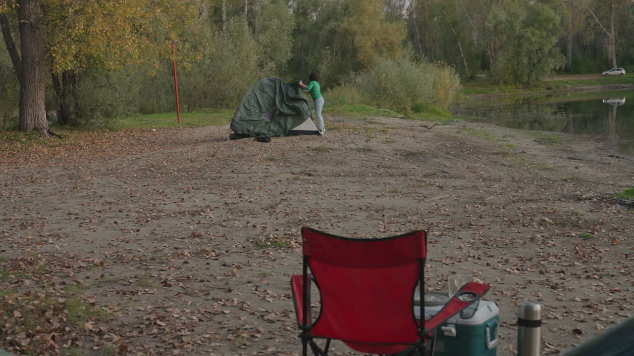 Hammock red chair tent on shore quiet lakeside camping scene lone green tent near tree line scattered autumn leaves on pebble beach empty hammock in foreground camping gear and thermos beside chair