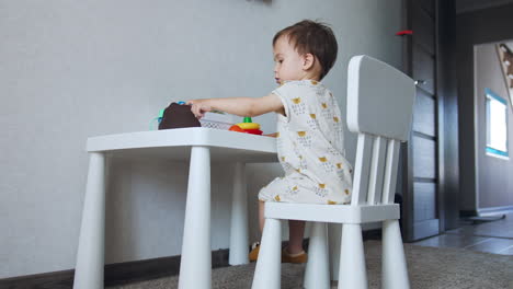toddler boy wearing white clothes sits at the desk. small kid plays bright toys. low angle view.
