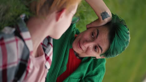 video vertical: una chica lesbiana con cabello corto verde en una camisa verde se apoya en su brazo y se comunica con su novia con una rubia con un corte de pelo corto en gafas en una camiseta rosa durante su cita en el parque acostada en el césped