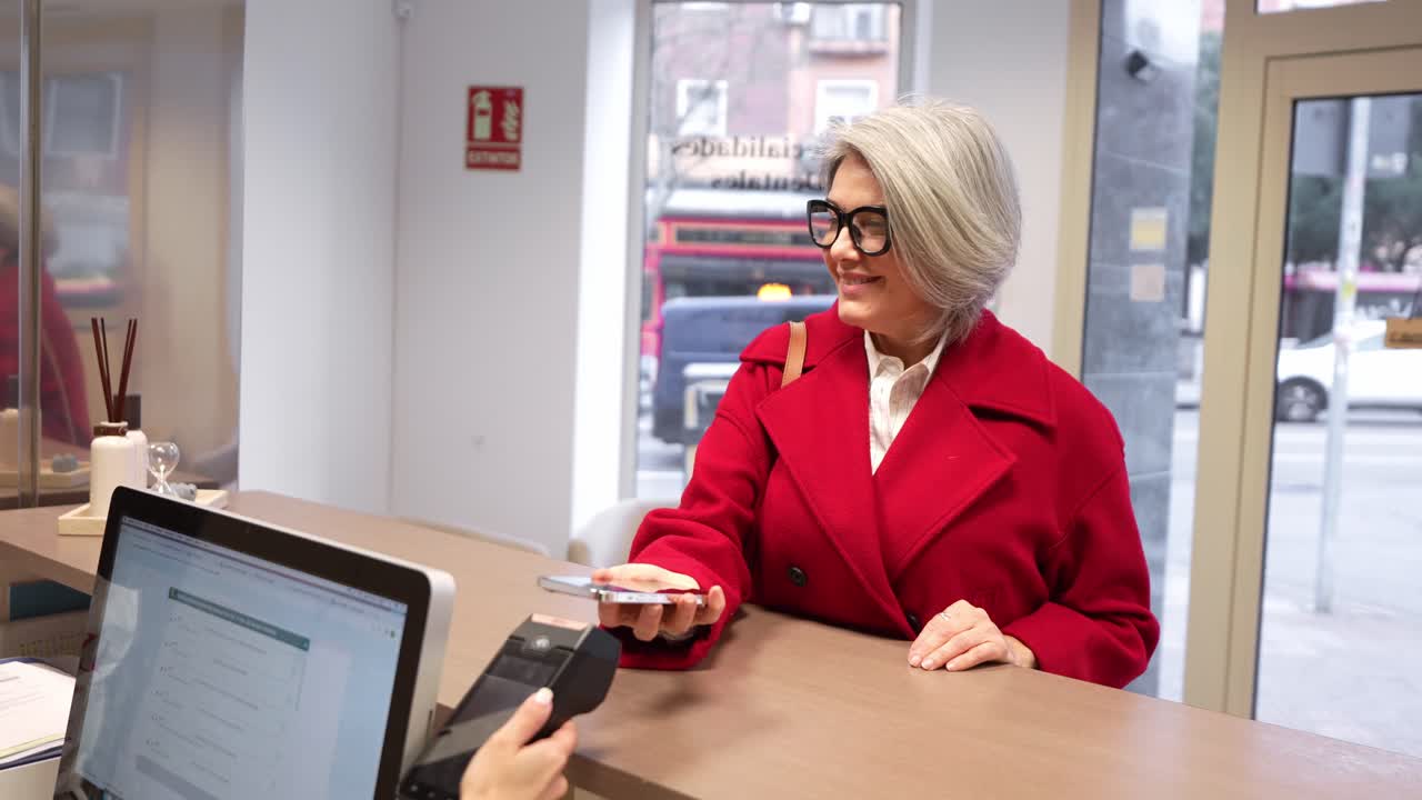 Reifere Frau tätigt kontaktlose Zahlung mit Smartphone an der Ladenkasse.