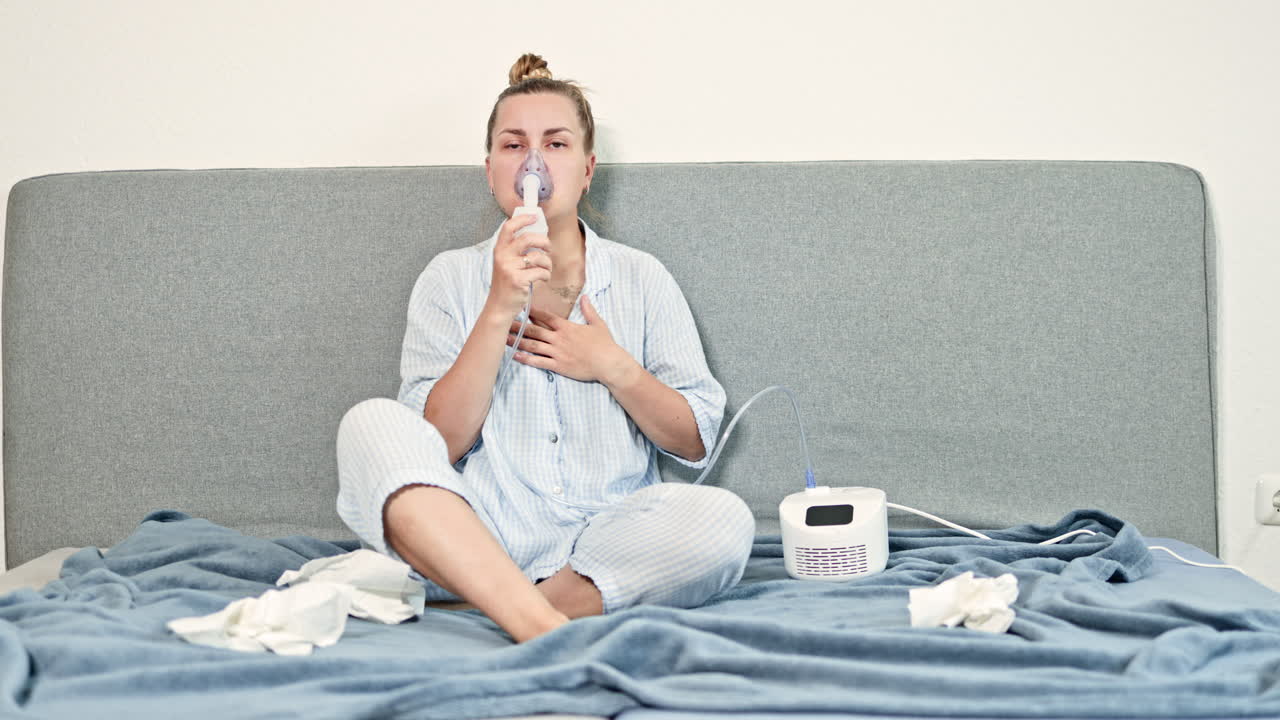 Giovane donna seduta sul letto che si mette una maschera sul viso per fare un'inalazione terapeutica. Donna che inala per il trattamento della tosse. Terapia con nebulizzatore a casa. Autotrattamento con soluzione fisiologica e inalatore.