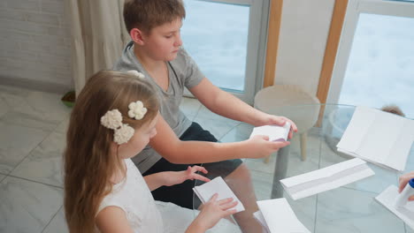 kids focused on creating paper craft, folding paper carefully at table, clear concentration on their hands as they work, surrounded by other folded papers in a cozy home environment