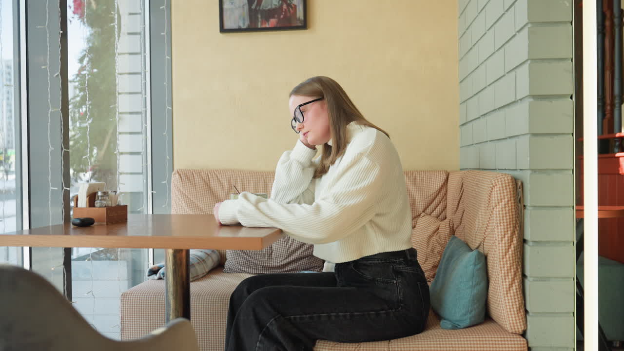 Woman seated on checkered bench stirs tea thoughtfully near large window, with soft daylight casting shadows on cozy cafe interior featuring framed wall art, pillows, and hanging light fixture