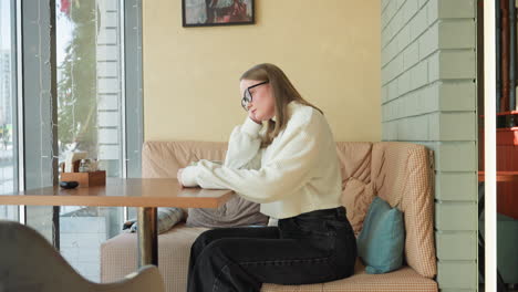 woman seated on checkered bench stirs tea thoughtfully near large window, with soft daylight casting shadows on cozy cafe interior featuring framed wall art, pillows, and hanging light fixture