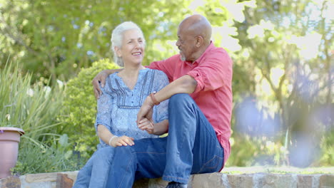 Feliz-Pareja-Birracial-Senior-Sentada-En-La-Pared-Y-Hablando-En-El-Soleado-Jardín-De-Casa,-Cámara-Lenta