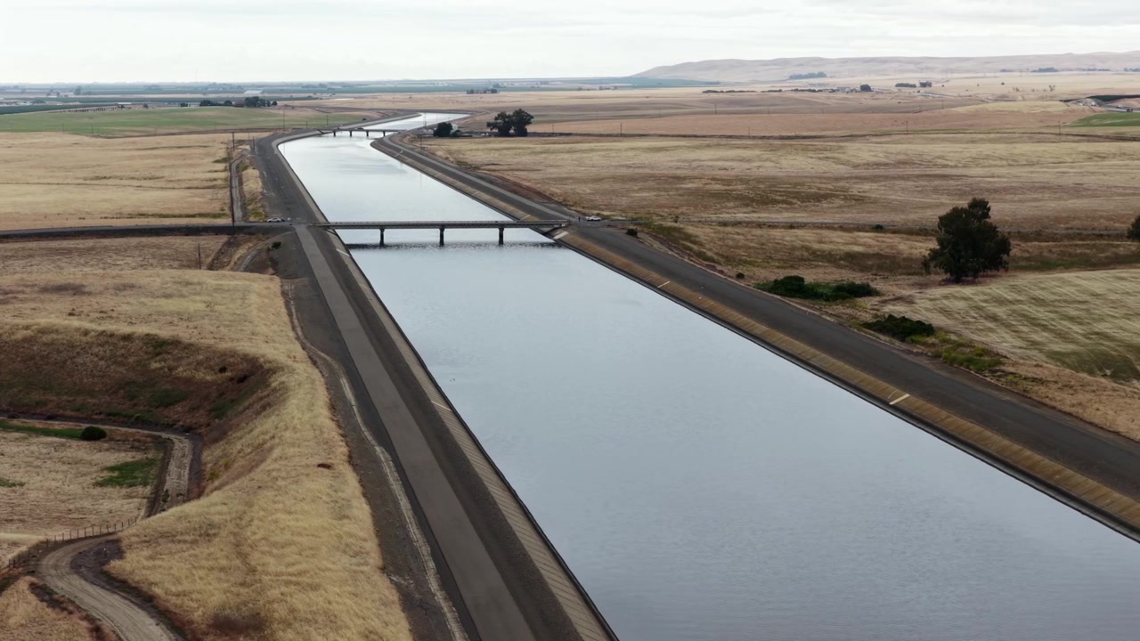 Das California Aquädukt schneidet geradewegs durch trockene goldene Felder, eine Straßenbrücke überquert auf halbem Weg, mit flachem Tal und entfernten dunstigen Hügeln unter blassem bedecktem Himmel. Kalifornien, USA. Luftaufnahme.