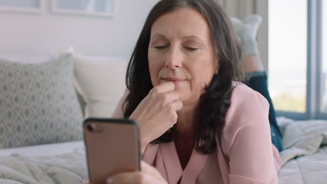 Mujer-Madura-Feliz-Usando-Un-Teléfono-Inteligente-Relajándose-En-La-Cama-Disfrutando-Navegando-Mensajes-De-Texto-En-Línea-Compartiendo-Su-Estilo-De-Vida