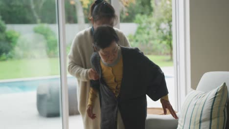 Ältere-Asiatische-Mutter-Wird-Von-Erwachsener-Tochter-Im-Wohnzimmer-In-Den-Sessel-Geholfen
