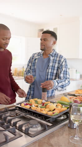 vertical video of african american gay male couple seasoning vegetables in kitchen, slow motion
