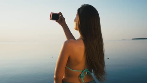 una joven bonita tomando fotos con su teléfono inteligente al atardecer en la playa cerca del mar en vacaciones. cámara lenta